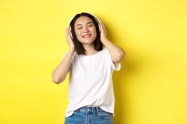 Mulher asiática atraente, curtindo ouvir música em fones de ouvido sem fio, sorrindo, satisfeita e dançando com os olhos fechados, em pé sobre um fundo amarelo.