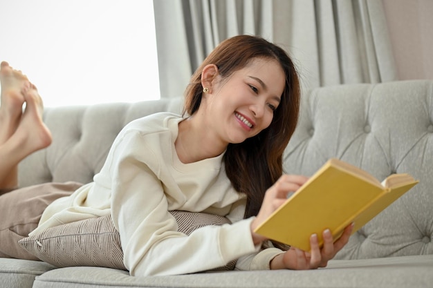 Mulher asiática atraente deitada em um sofá na sala de estar gosta de ler um livro de ficção de aventura
