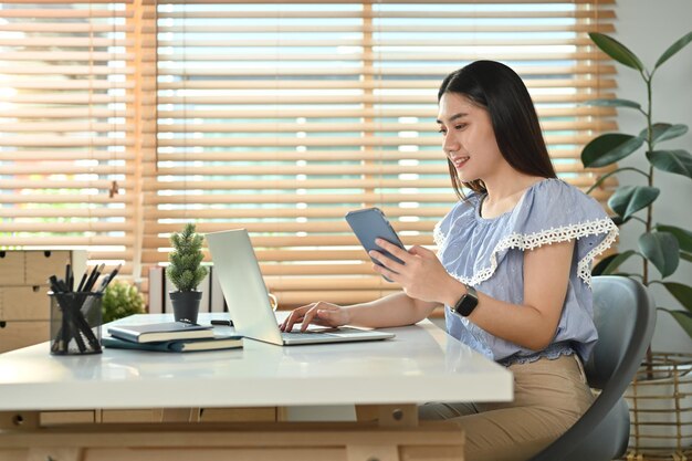 Mulher asiática atraente freelancer segurando telefone inteligente e usando laptop no escritório doméstico moderno