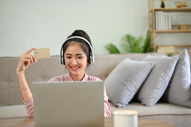 Mulher asiática atraente olhando para a tela do laptop segurando um cartão de crédito em sua sala de estar