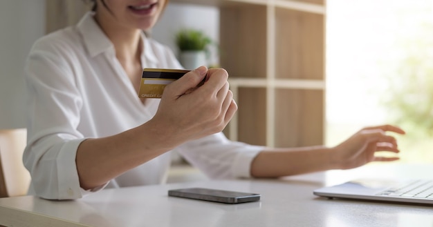 Mulher asiática atraente sentada relaxada no sofá, segurando um cartão de crédito e smartphone, provavelmente comprando algo online.