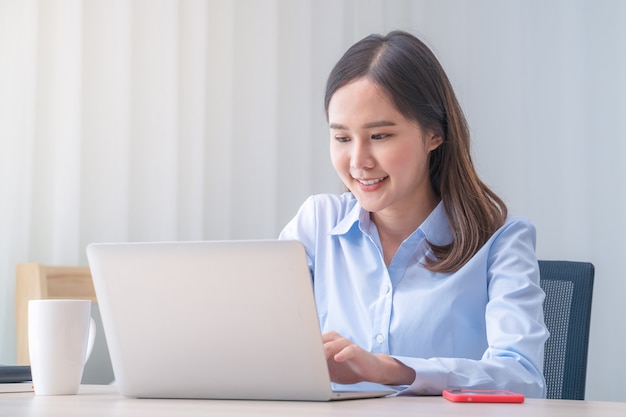 Mulher asiática atraente trabalha em casa com o laptop na mesa no quarto. moça que sorri com cara da felicidade ao ler na tela de computador. teletrabalho, freelancer e conceito de estilo de vida moderno.