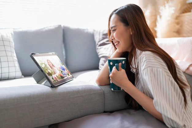 Mulher asiática atraente usa pano casual em casa isolar quarentena gosta de assistir streaming bradcast programa de filmes on-line via tablet no chão ao lado do sofá na sala de estar interior aconchegante fundo