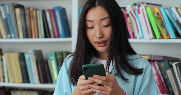 Mulher asiática atraente usando mensagem de texto de smartphone em pé na biblioteca