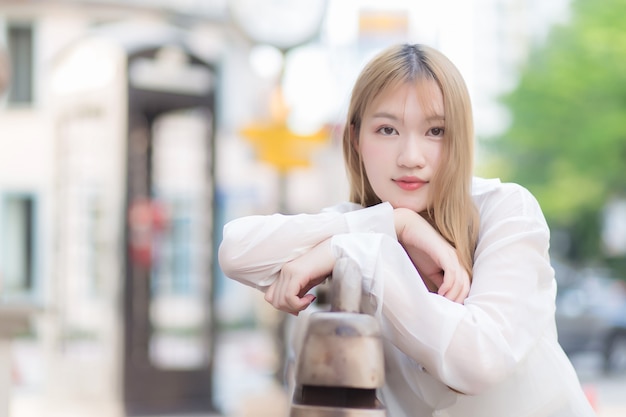 Mulher asiática bonita com cabelo cor de bronze na camisa branca enquanto se senta feliz na beira da rua da cidade
