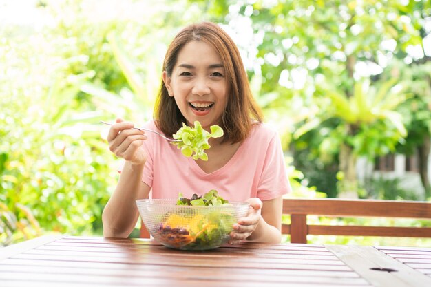 Foto mulher asiática bonita e saudável feliz sentada no terraço e comendo uma salada verde saudável
