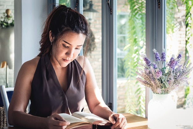 Mulher asiática bonita nova que senta-se na tabela e que lê livro interessante