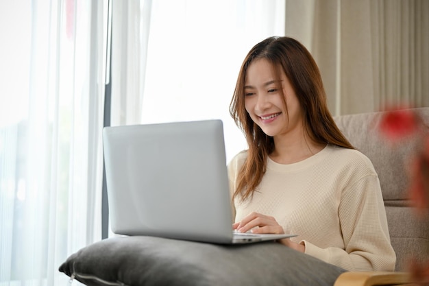 Mulher asiática bonita relaxa na sala de estar usando um laptop navegando na internet