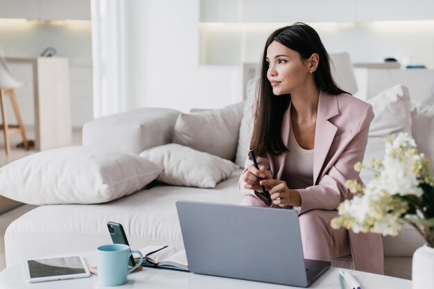 Mulher asiática bonita séria em terno de negócio sentado no sofá aconchegante na mesa com telefone portátil xícara de café em casa trabalhando remotamente Empresário atraente segurando a caneta olhando de lado Pessoas de negócios