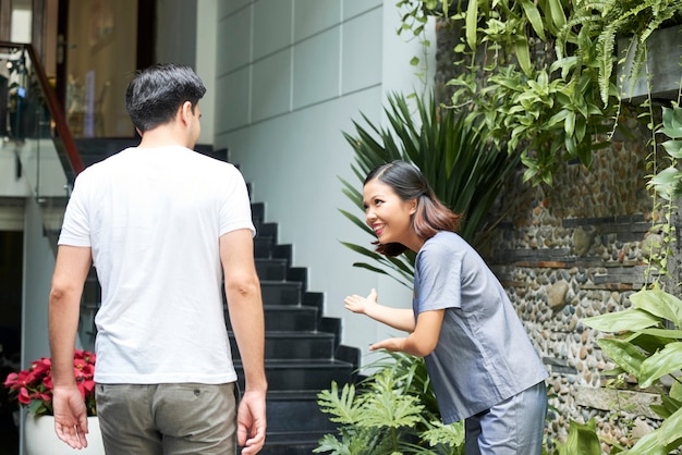 Mulher asiática bonita sorridente fazendo uma reverência para um homem entrando no prédio do salão de spa e fazendo um gesto convidativo