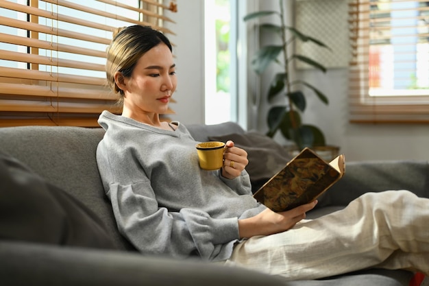 Mulher asiática calma bebendo chá quente de ervas e lendo livro no sofá em casa