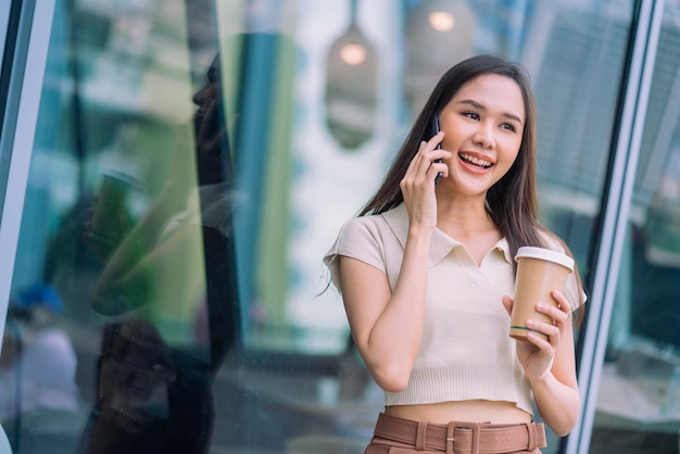 Mulher asiática casual relaxa programador nômade digital parece conversa no smartphone enquanto caminha na cidade com uma xícara de café segurando um sentimento positivo alegre e sorridente, reduzindo o estilo de vida