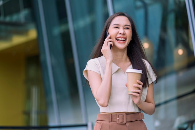 Mulher asiática casual relaxa programador nômade digital parece conversa no smartphone enquanto caminha na cidade com uma xícara de café segurando um sentimento positivo alegre e sorridente, reduzindo o estilo de vida