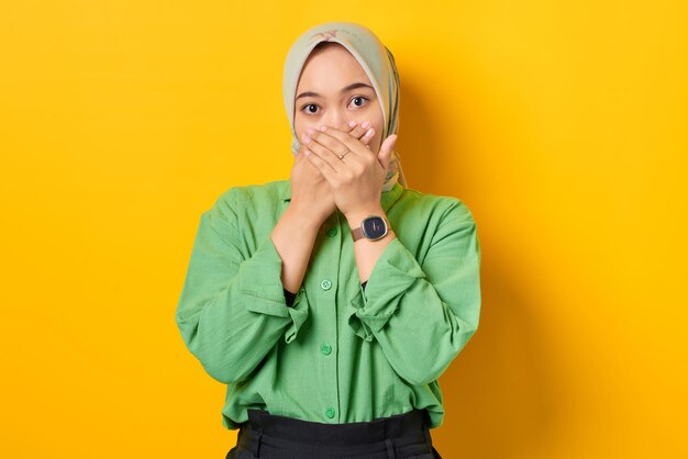 Mulher asiática chocada com camisa verde cobrindo a boca com ambas as mãos em fundo amarelo