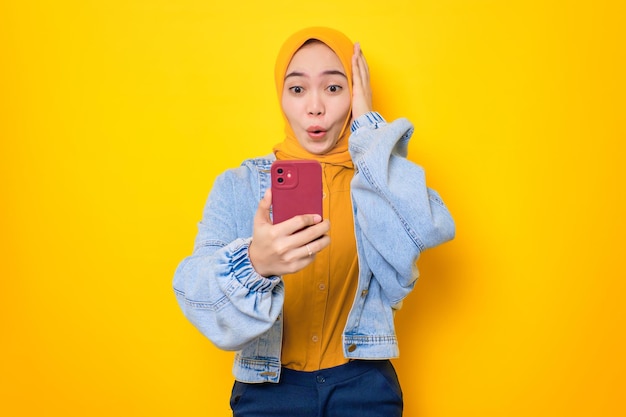 Mulher asiática chocada com jaqueta jeans olhando para a tela do celular com a boca aberta reagindo a ótimas notícias isoladas sobre fundo amarelo