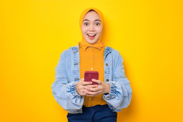 Mulher asiática chocada em jaqueta jeans segurando telefone celular e olhando para câmera isolada sobre fundo amarelo