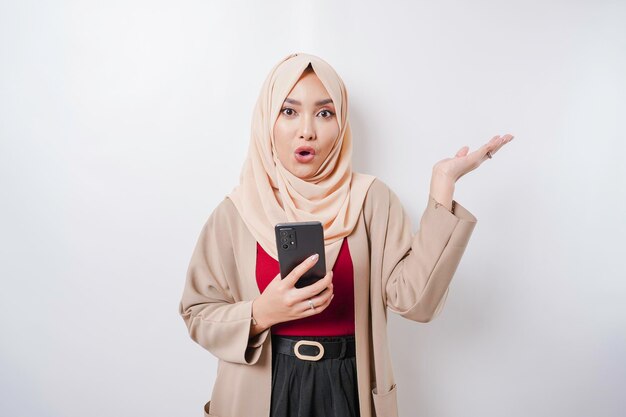 Mulher asiática chocada usando hijab apontando para o espaço da cópia ao lado dela enquanto segura o telefone isolado pelo fundo branco