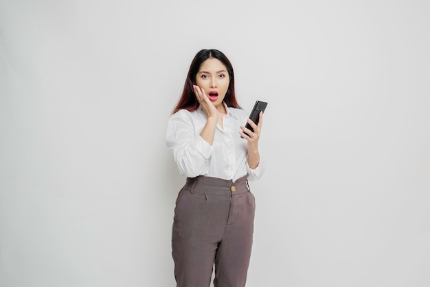 Mulher asiática chocada vestindo camisa branca e segurando seu telefone isolado por fundo branco