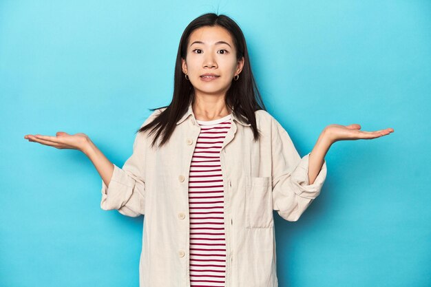 Mulher asiática com camisa em camadas e camiseta listrada, confusa e duvidosa, encolhendo os ombros