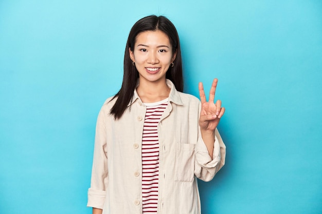 Mulher asiática com camisa em camadas e camiseta listrada mostrando sinal de vitória e sorrindo amplamente