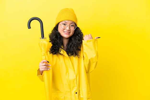 Foto mulher asiática com casaco à prova de chuva e guarda-chuva isolado em fundo amarelo apontando para o lado para apresentar um produto