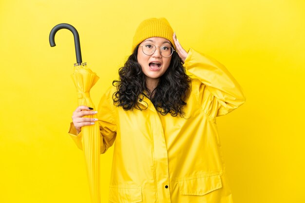 Mulher asiática com casaco à prova de chuva e guarda-chuva isolado em um fundo amarelo com expressão de surpresa