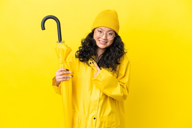 Mulher asiática com casaco à prova de chuva e guarda-chuva isolado em um fundo amarelo com expressão facial surpresa