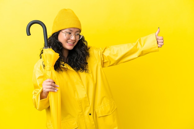 Foto mulher asiática com casaco à prova de chuva e guarda-chuva isolado em um fundo amarelo fazendo um gesto de polegar para cima