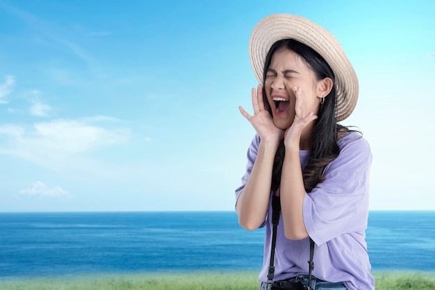 Mulher asiática com chapéu e câmera gritando algo com fundo de céu azul