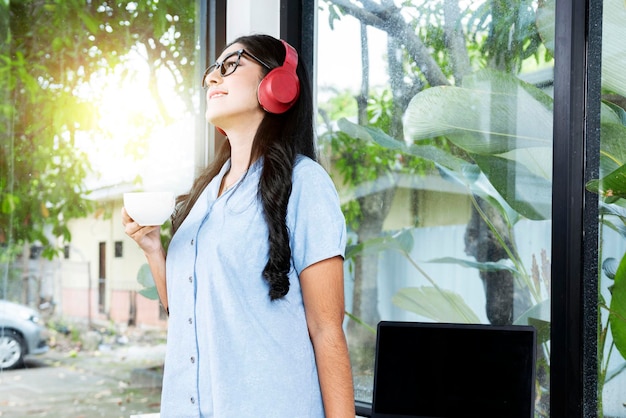 Mulher asiática com óculos ouvindo música com fones de ouvido e segurando uma xícara de café com um laptop na mesa