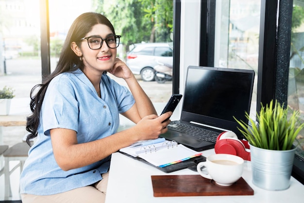 Mulher asiática com óculos usando um telefone celular com um laptop e um notebook com uma xícara de café na mesa