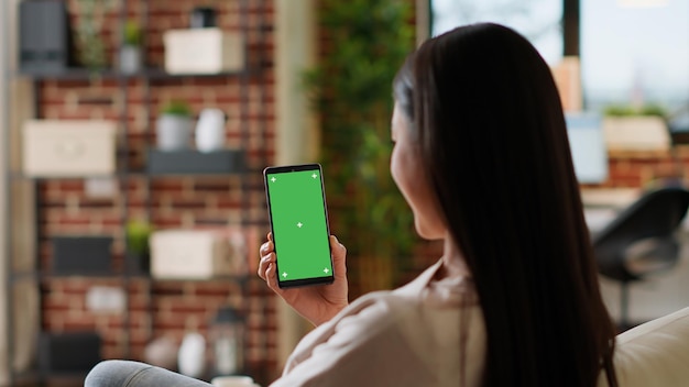 Mulher asiática com smartphone com tela verde enquanto está sentado em casa no sofá. Adulto jovem sentado no sofá com celular móvel com fundo de croma de maquete.