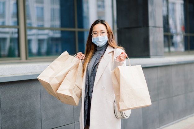 Mulher asiática com um casaco, óculos e uma máscara médica fica na rua, segurando um pacote nas mãos.