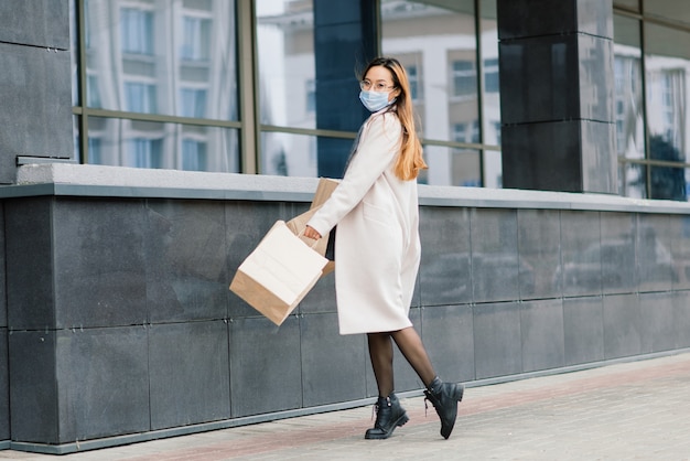 Mulher asiática com um casaco, óculos e uma máscara médica fica na rua, segurando um pacote nas mãos.
