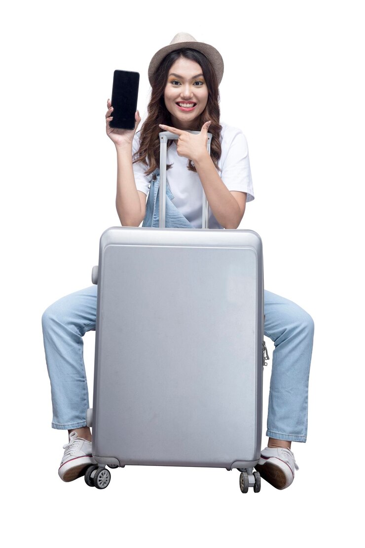 Foto mulher asiática com um chapéu sentado em uma mala e mostrando uma tela de celular vazia