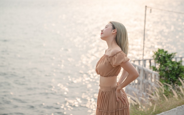 Mulher asiática com vestido posando na praia, momento do sol dourado do mar.