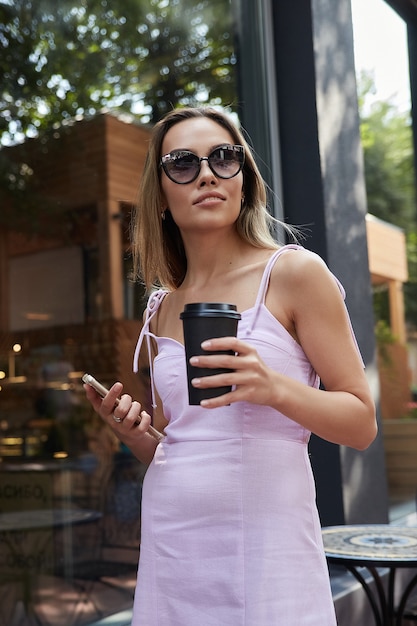 Mulher asiática com vestido rosa em pé ao ar livre segurando um smartphone e uma xícara de café