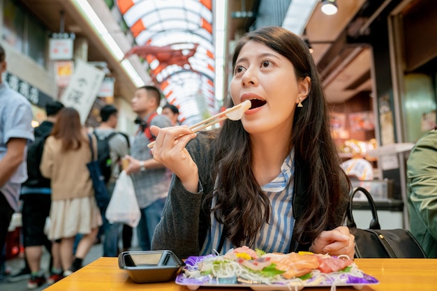 mulher asiática comendo sashimi no mercado. um prato de peixe cru e vista da multidão.