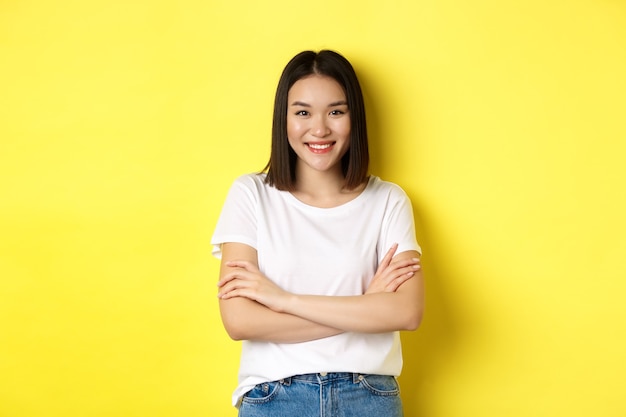 Mulher asiática confiante e elegante com os braços cruzados no peito e sorrindo, em pé sobre um fundo amarelo.