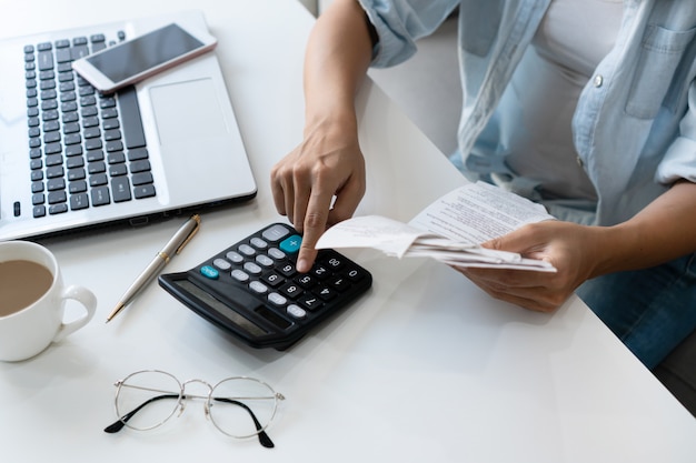 Mulher asiática consideravelmente nova que usa a calculadora ao guardar contas para calcular despesas e impostos home na sala de visitas em casa.