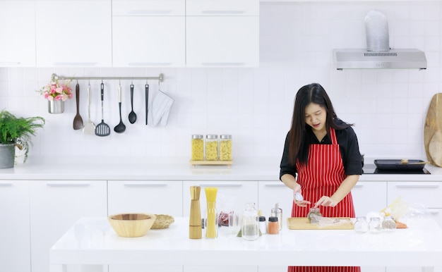 Mulher asiática contente no avental de pé à mesa com ingredientes variados e preparando comida saborosa na cozinha em casa.
