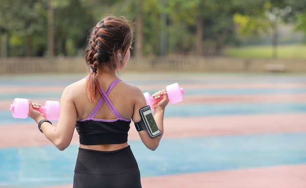 Mulher asiática de beleza jovem atlética vista traseira no sportswear em pé e levantando halteres ao ar livre para o exercício matinal. garota ativa malhar em exercícios de bíceps com fundo de árvore verde. conceito de esporte