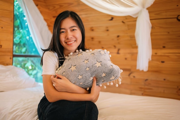 Foto mulher asiática de cabelos pretos sentada em uma cama branca em um quarto decorado em madeira marrom abraçando uma almofada