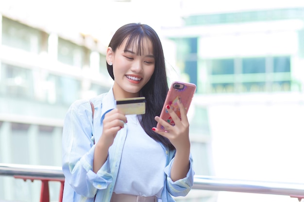 Mulher asiática de camisa azul sorri com sucesso ao ar livre na cidade enquanto segura smartphone e cartão de crédito