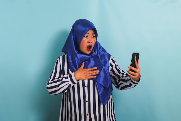 Mulher asiática de meia-idade chocada em hijab está olhando para o telefone isolado em um fundo azul