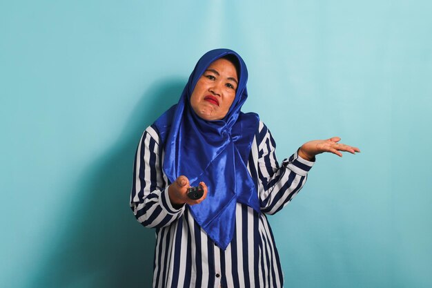 Mulher asiática de meia-idade entediada em hijab está mudando de canal isolada em fundo azul