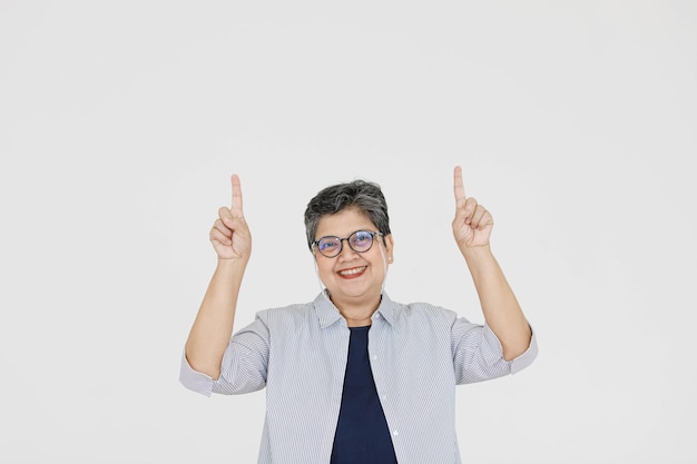 Mulher asiática de meia-idade otimista de óculos, olhando para a câmera com um sorriso e apontando para um fundo cinza