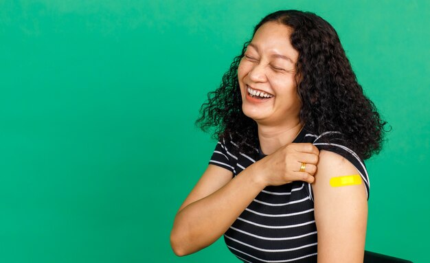 Foto mulher asiática de meia-idade segurando seu braço com curativo mostrando que ela foi vacinada contra o vírus covid 19 em fundo verde. conceito para vacinação contra covid 19.