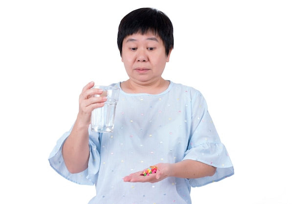 Mulher asiática de meia-idade segurando um copo d'água e olhando muitos comprimidos que precisa tomar isolados no fundo branco