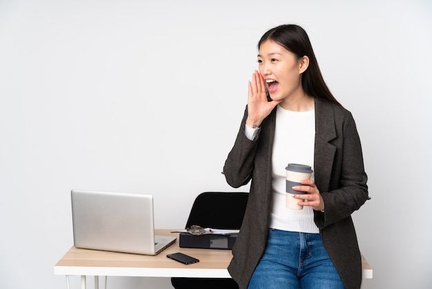 Mulher asiática de negócios em seu local de trabalho na parede branca, gritando com a boca aberta para a lateral